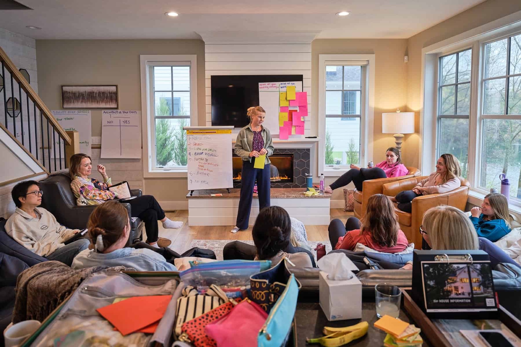 A group seated in a circle in a bright lakehouse living room, flip charts and sticky notes covering the wall behind a woman presenting.