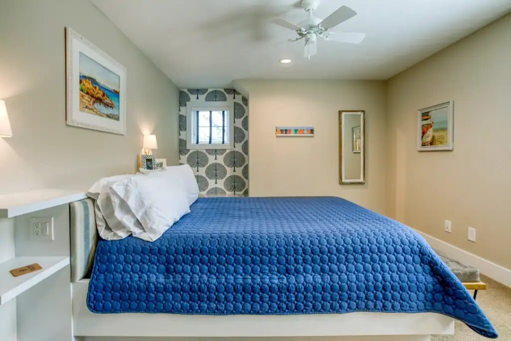 A bedroom with a large bed under a deep blue quilt, pale walls, patterned wallpaper behind the headboard and a bench at the foot of the bed.