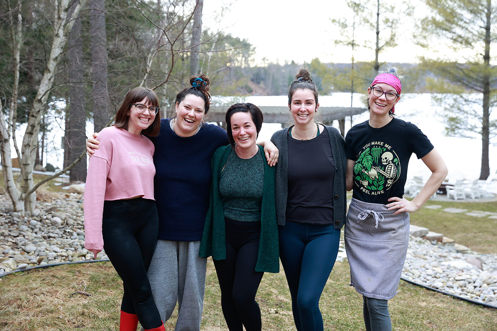 Five women standing arm in arm on the grass by a lake, laughing together, one still wearing an apron.