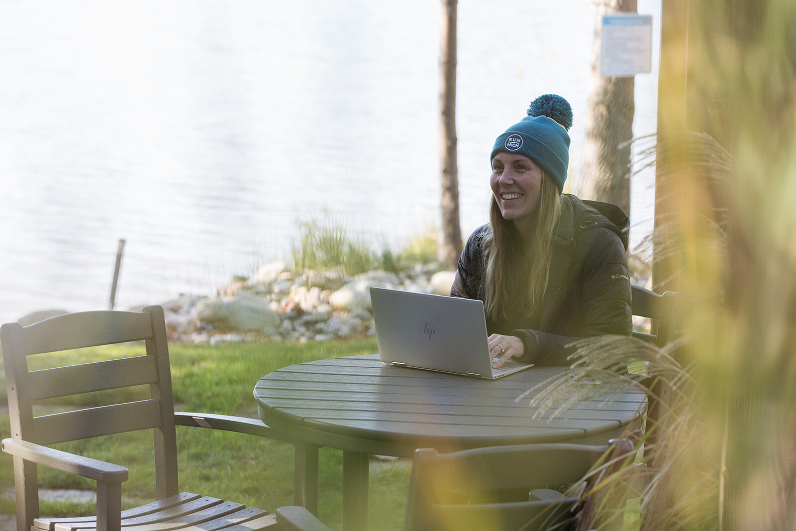 A woman working on a laptop at an outdoor table beside a lake, wrapped up against the cold.